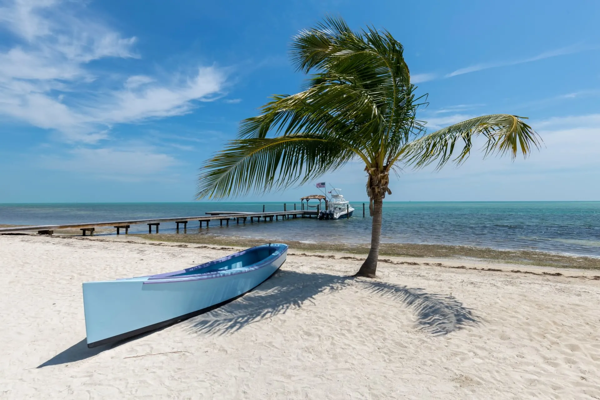 white sand beach with a blue canoe on it under a palm tree. There is a dock with a fishing boat in the background